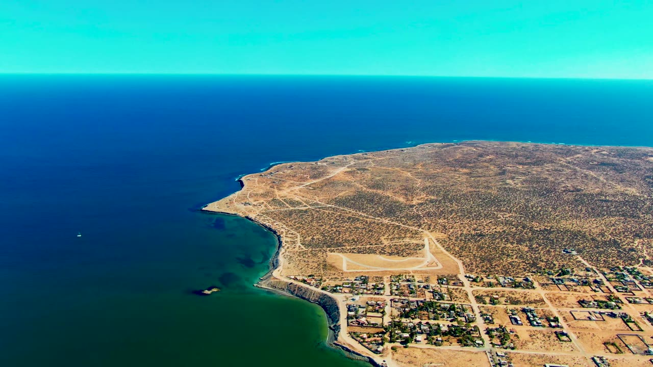 Opening aerial frame over San Juanico, Baja California Sur showing blue-green ocean beside a sandy coast, a small town grid, a pale runway, desert scrub, and no visible people.