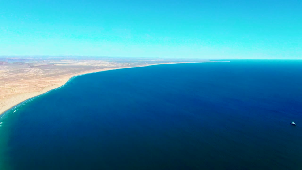 High aerial view over San Juanico, Baja California Sur, with sandy desert shoreline at left, deep blue water across the frame, a small boat near the lower right, and no visible people.