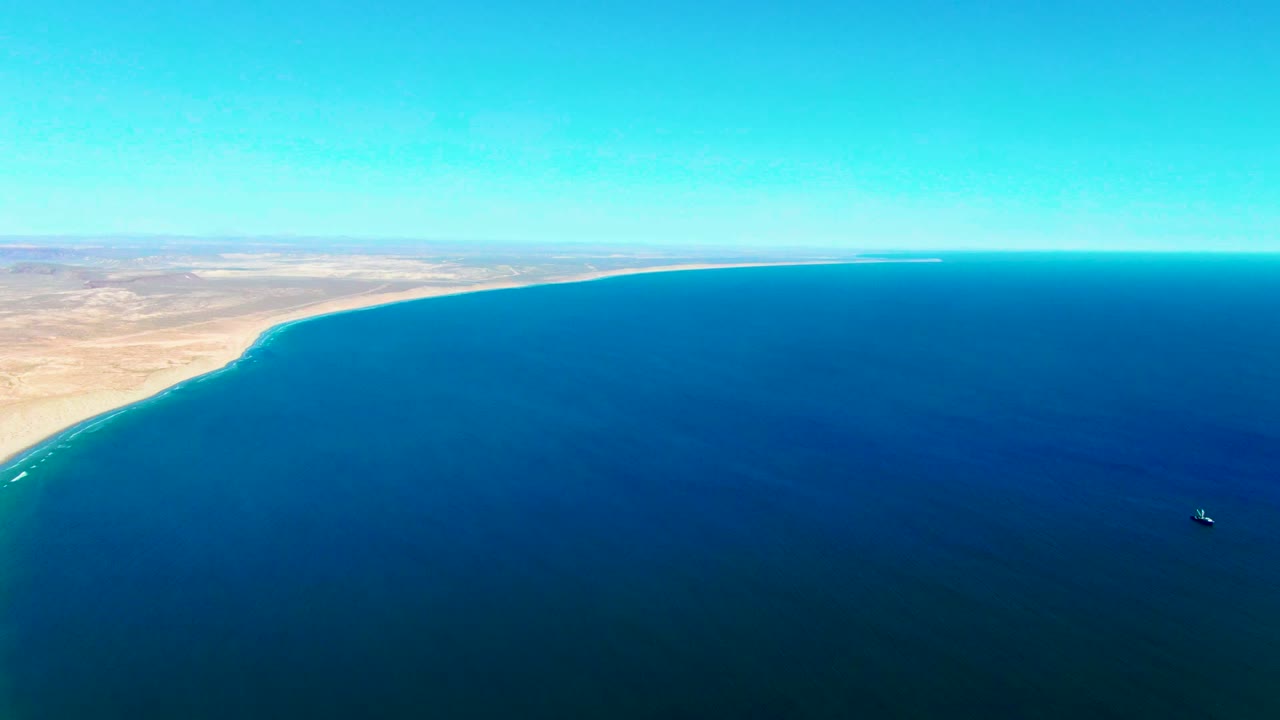 Wide aerial coastline with sandy land, clear sky, blue sea, and a small boat offshore.