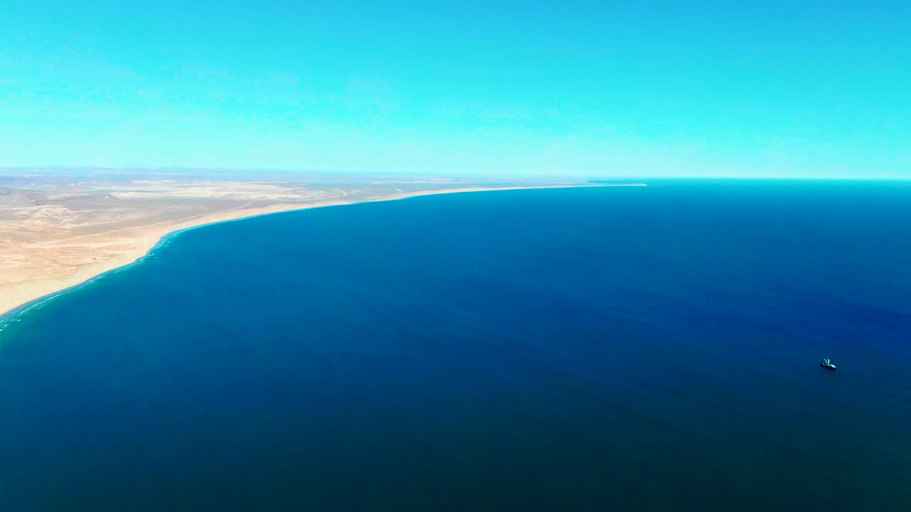 Wide drone-style view of San Juanico coastline, with pale desert land along the left and top edge, darker blue ocean filling the foreground, a small boat to the right, and no visible people.