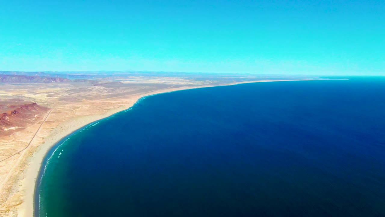 Aerial view over San Juanico, Baja California Sur, with a sandy desert coast on the left, deep blue ocean on the right, distant mountains, clear sky, and no visible people.