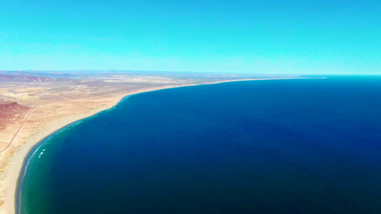 Aerial coastal view in San Juanico, Baja California Sur, with the shoreline running diagonally along the left edge, small white surf marks near the beach, open ocean across the frame, and no visible people.