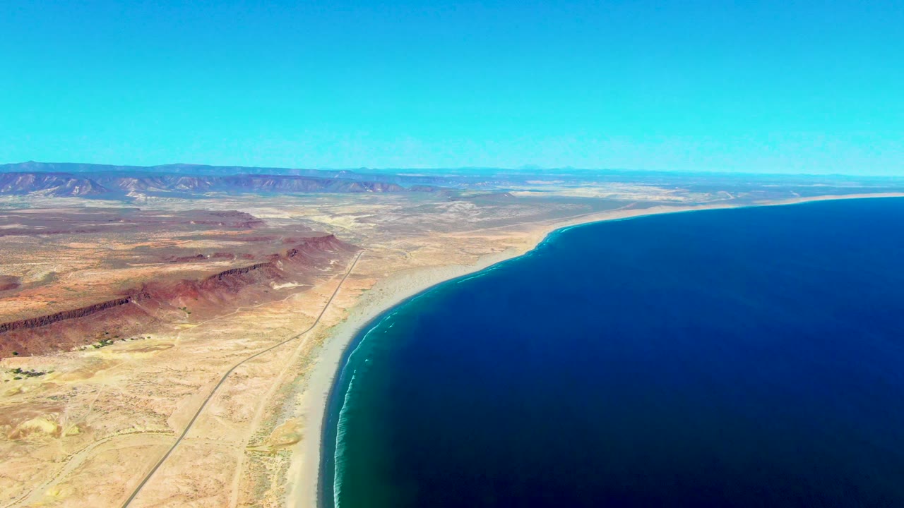 Opening aerial frame over San Juanico, Baja California Sur, with deep blue ocean on the right, a curved sandy beach, desert road and hills on the left, distant mountains, clear sky, and no visible people.