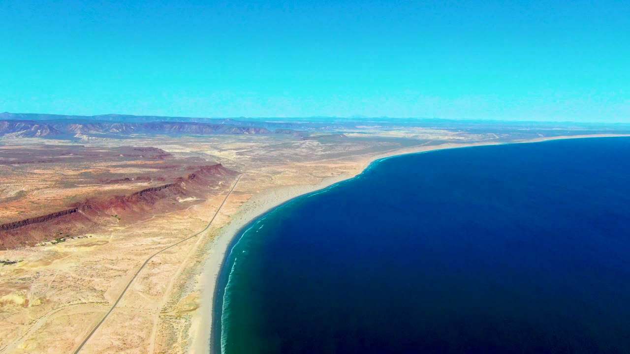 Aerial coastline view with ocean to the right and desert road and mountains to the left.