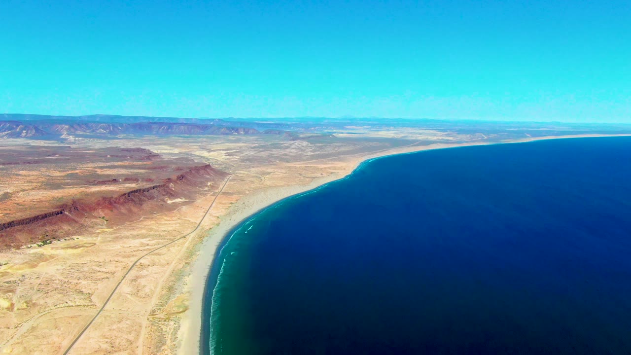 Third frame presents San Juanico's curved coast with a wider sweep of dark blue ocean, tan desert flats, a visible road near the beach, distant mountains under clear sky, and no visible people.