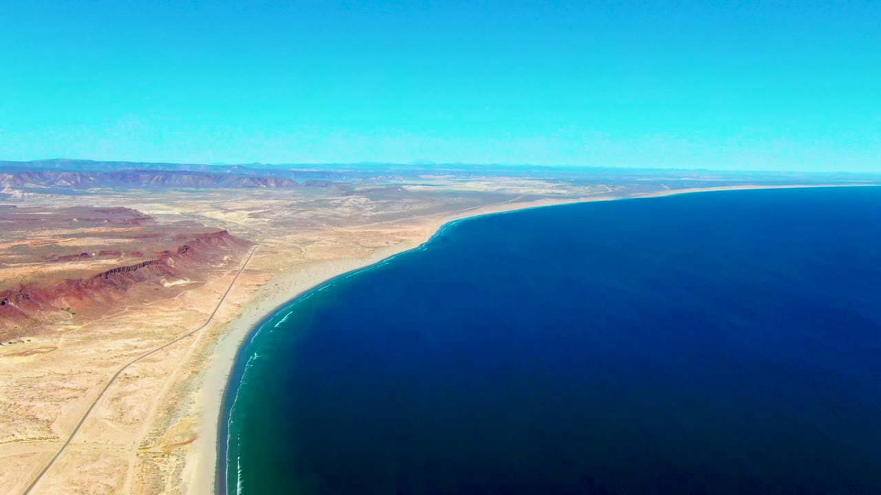 Aerial view of San Juanico, Baja California Sur coastline with tan desert, a pale beach, deep blue ocean, clear sky, and no visible people.