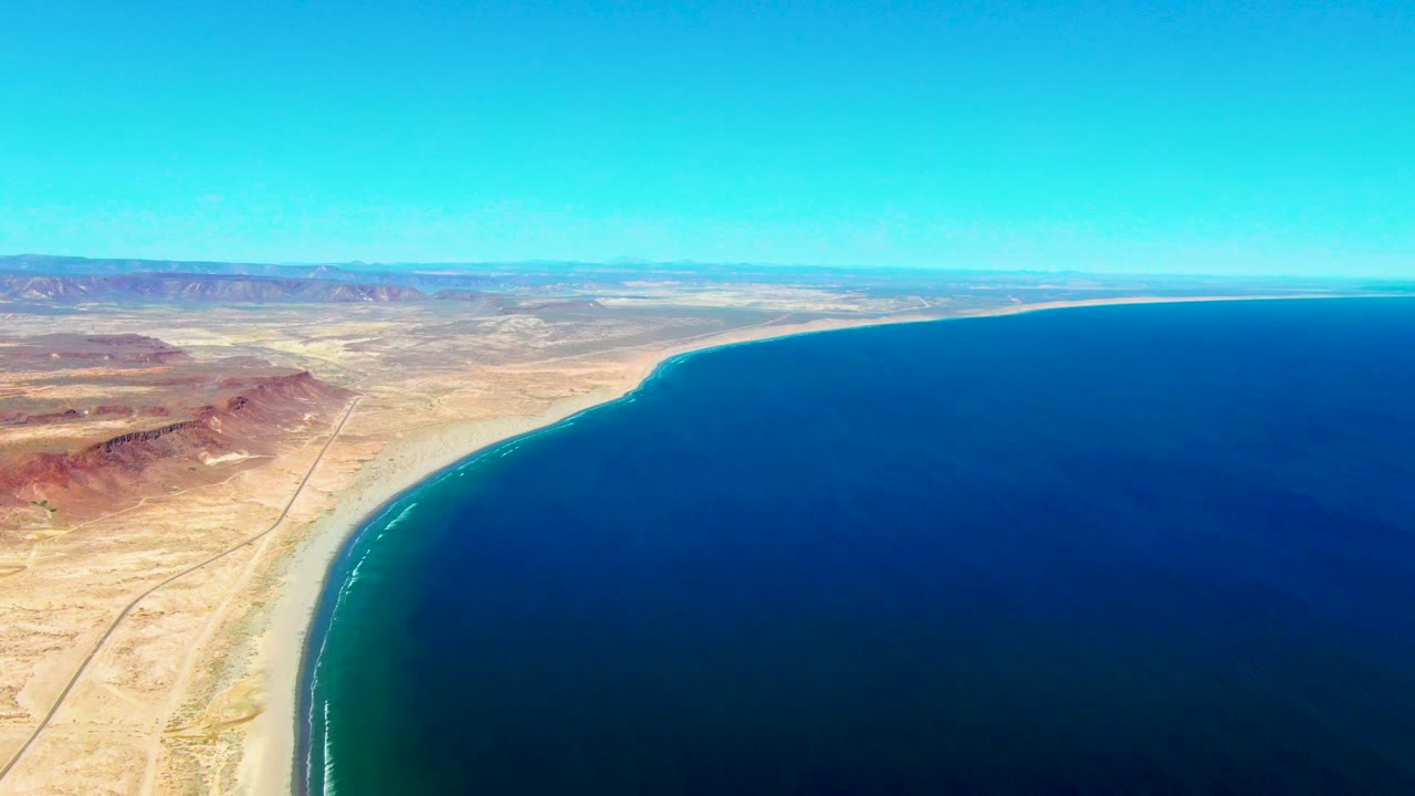 Wide aerial view over San Juanico with reddish desert hills to the left, sandy coast below, open blue water to the right, and no visible people.