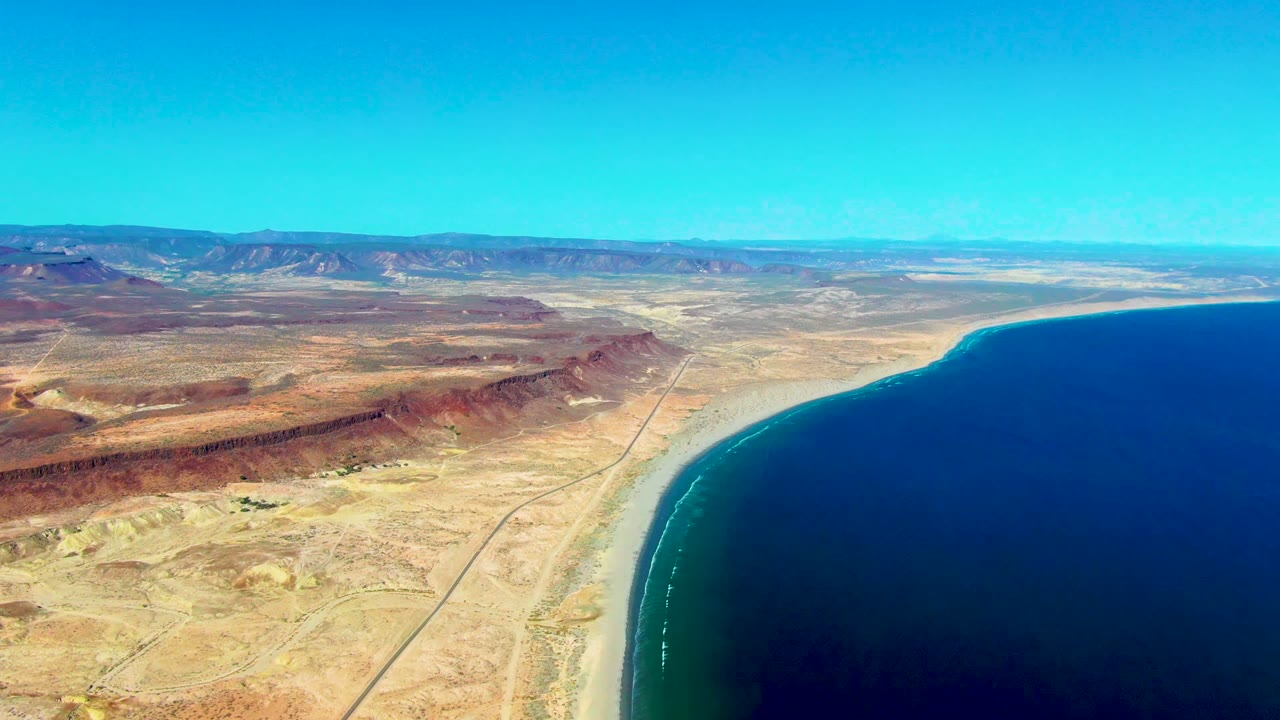 Opening aerial frame over San Juanico, Baja California Sur, with tan desert, a straight coastal road, pale beach, deep blue ocean, distant ridges, and no visible people.