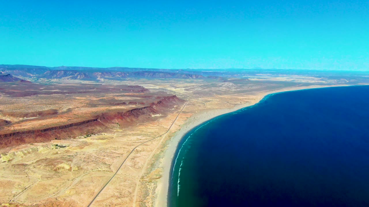 Drone-height view of San Juanico desert coast with reddish ridges left of frame, a pale road near the beach, dark blue water to the right, and no visible people.