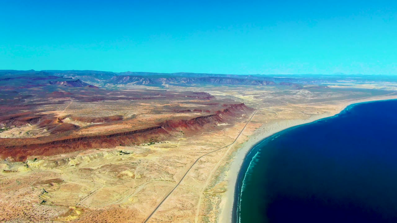 Aerial view over San Juanico, Baja California Sur, with tan desert, red-brown ridges, a straight road near the coast, blue ocean on the right, distant mountains, and no visible people.