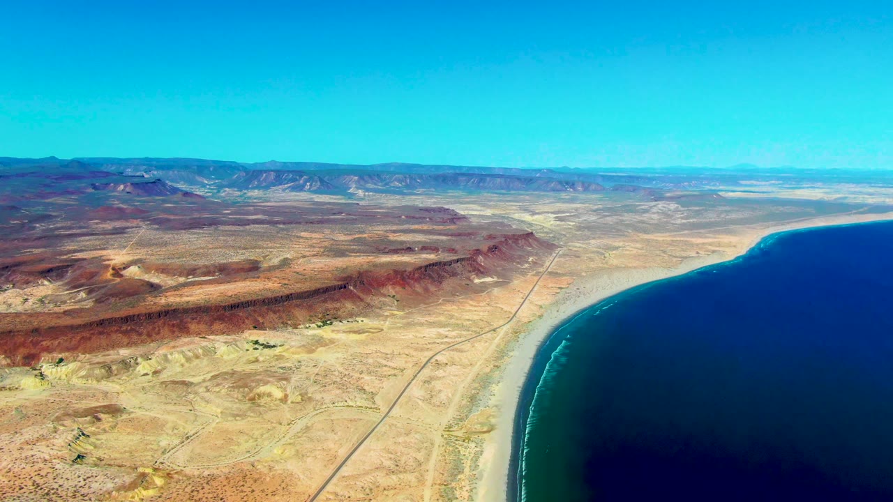 Wide aerial coastline with arid hills, road, and dark blue ocean