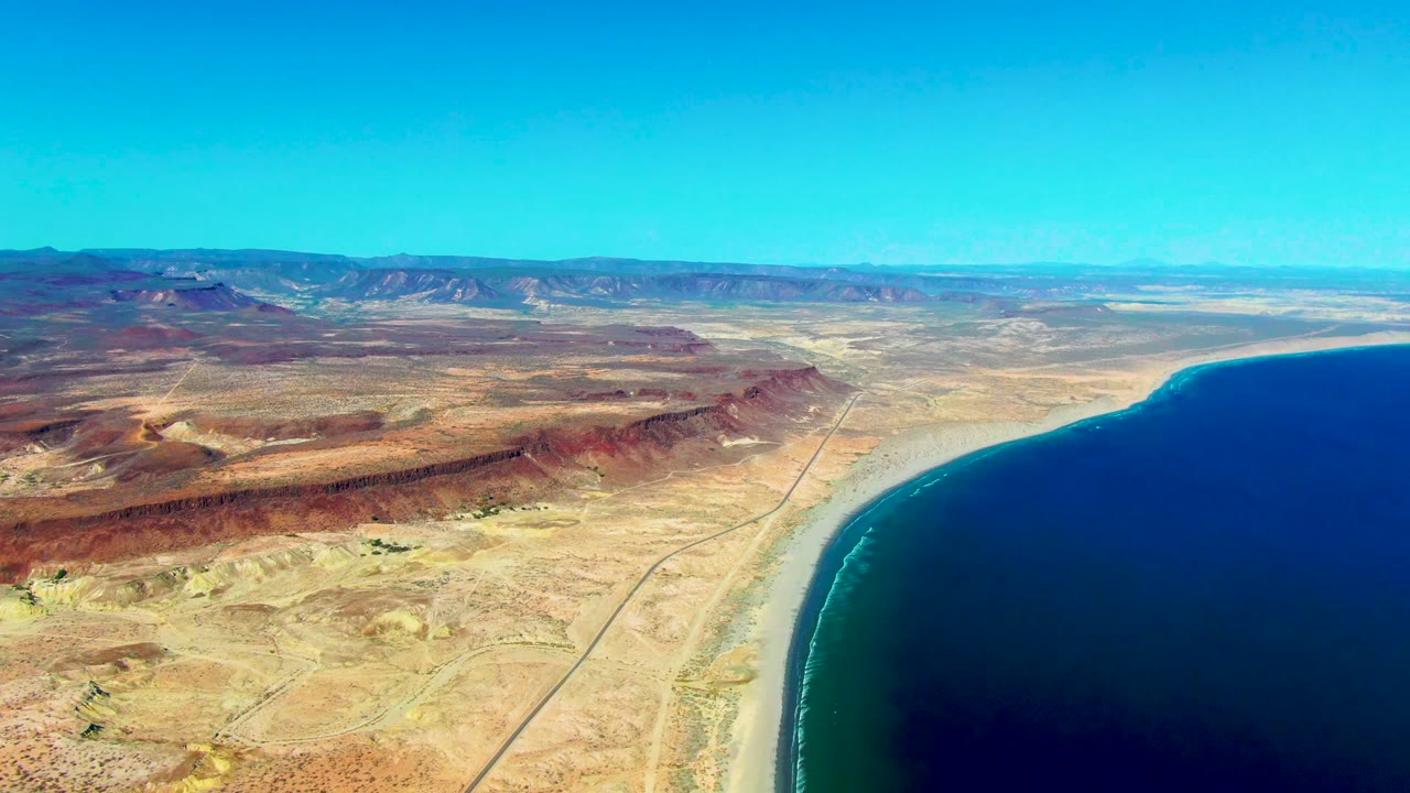 Aerial view in San Juanico, Baja California Sur, showing the road nearly parallel to the pale shoreline, red and tan desert land to the left, deep ocean to the right, distant mountains, and no visible people.