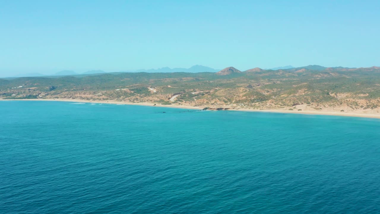 Aerial view at El Tule Los Cabos showing turquoise ocean in the foreground, a sandy shoreline, dry low hills, hazy mountains, clear sky, and no visible people.