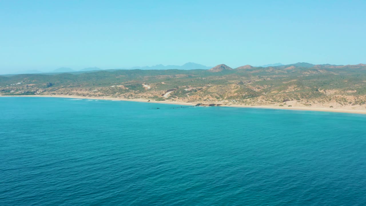 Aerial coastline with calm blue water, pale sand, and dry hills beyond.