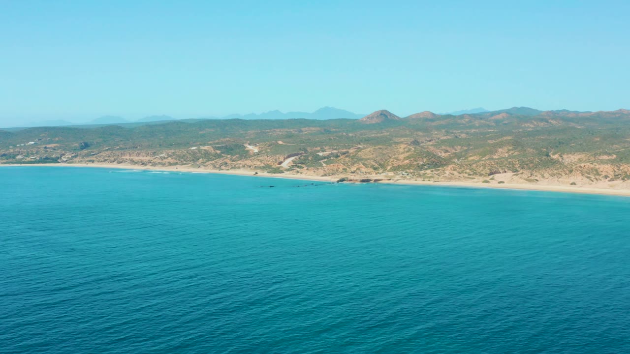 Aerial view over El Tule Los Cabos with rippled blue ocean, a long sandy beach, scrubby low hills, faint mountains, clear sky, and no visible people.