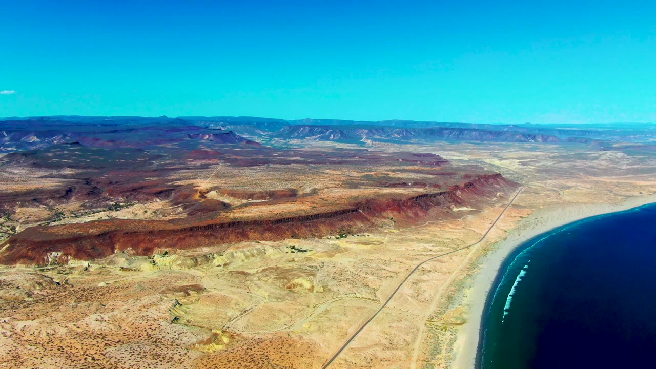 Aerial frame over San Juanico, Baja California Sur showing tan desert terrain, rust ridges, a straight coastal road, dark blue ocean at right, distant mountains, clear sky, and no visible people.