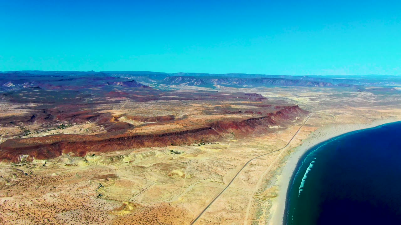 High aerial coastline with desert ridges, beach, road, and blue water.