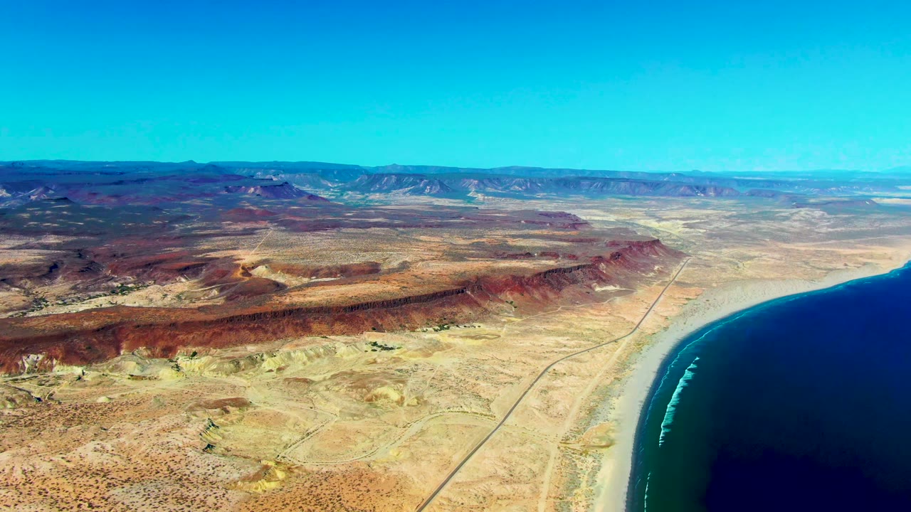 Aerial view over San Juanico, Baja California Sur where the dark ocean occupies the right edge, a sandy beach and straight road trace the shore, and dry desert flats lead to a long mountain horizon with no visible people.