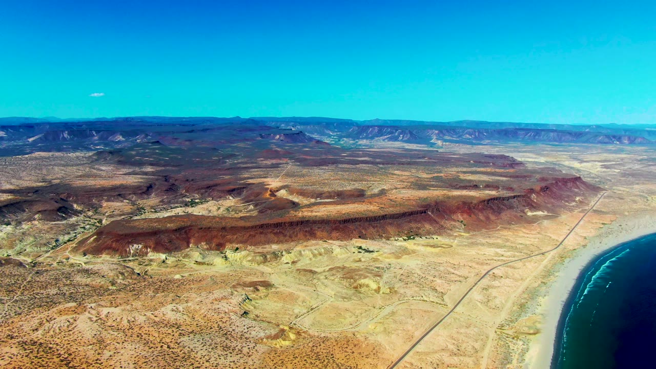 Aerial frame over San Juanico, Baja California Sur showing tan desert terrain, red-brown ridges, a straight road, blue ocean at right, distant mountains, clear sky, and no visible people.