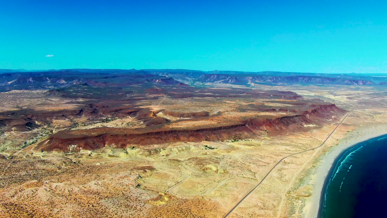 Wide aerial of sandy desert, red ridges, coastal road, ocean edge, and distant mountains.