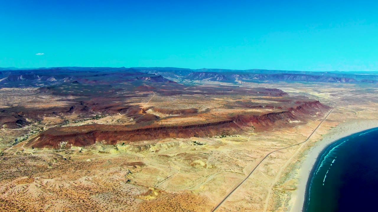At two seconds, the San Juanico coastline remains wide, with white surf lines visible in the blue water, a straight road inland, layered desert ridges, and no visible people.