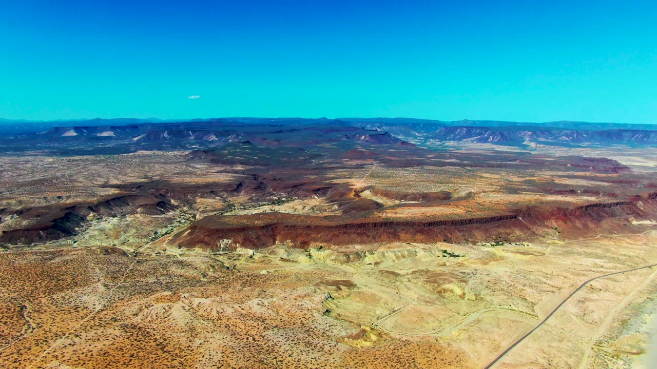 Wide aerial view over San Juanico, Baja California Sur desert with red ridges, pale flats, distant mountains, and no visible people.
