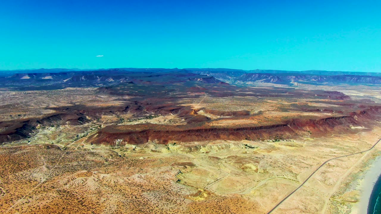 High drone view in San Juanico showing pale desert ground, a straight road line, reddish hills, distant mountains, and no visible people.