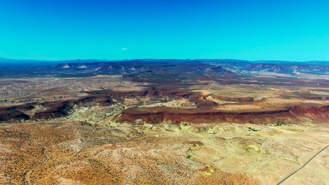 Opening aerial frame over San Juanico, Baja California Sur desert with tan flats, rust mesa bands, distant mountains, clear blue sky, and no visible people.