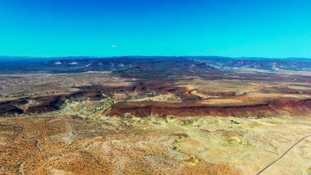 Wide desert aerial with mesa ridge, pale washes, scrub, and distant plateau