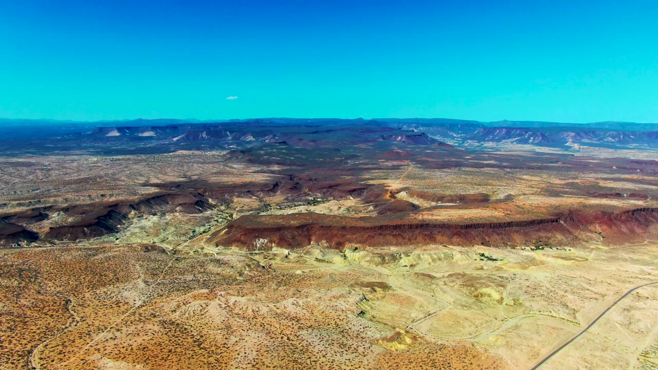 Mid-clip aerial frame in San Juanico, Baja California Sur with eroded pale ridges, low scrub, a faint straight road trace, distant mesas, and no visible people.