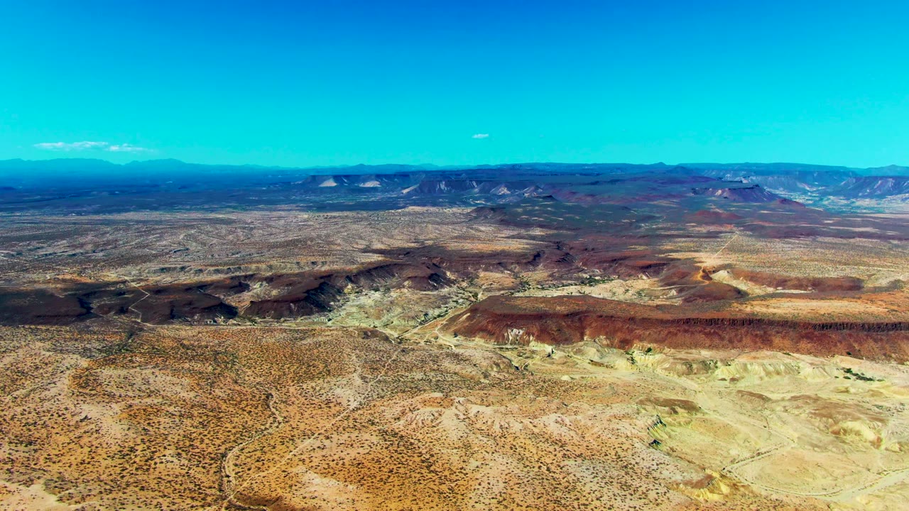 Opening frame of San Juanico, Baja California Sur desert terrain with tan scrub, red-brown mesas, distant ridges, a small white cloud, and no visible people.