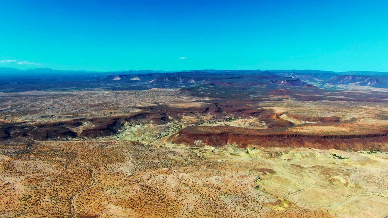 Opening frame over San Juanico, Baja California Sur, showing a wide desert plateau, red-brown mesas, pale washes, distant ridgelines, clear blue sky, and no visible people.