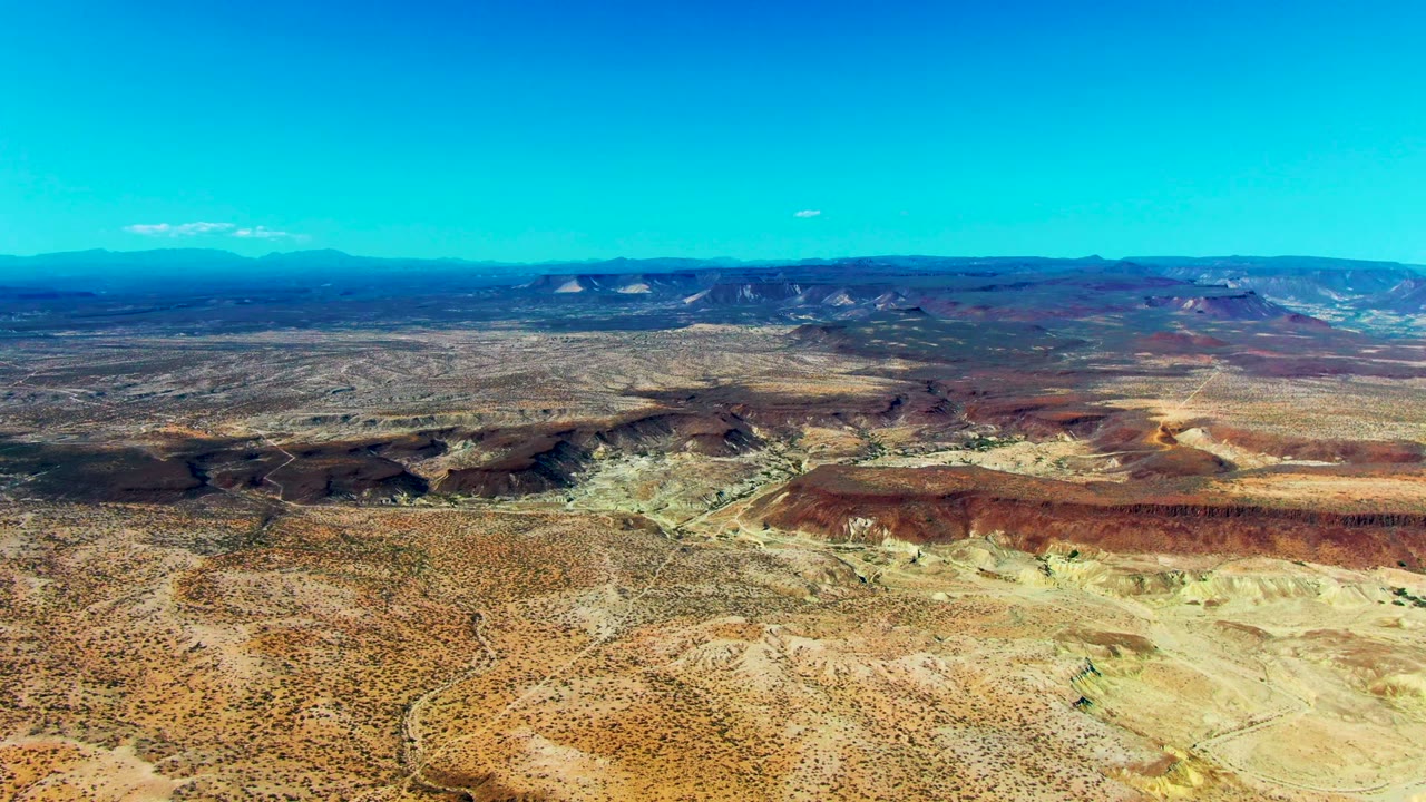 San Juanico desert aerial with red and orange rock formations crossing the middle of frame, pale eroded ground below, distant mountains, and no visible people.