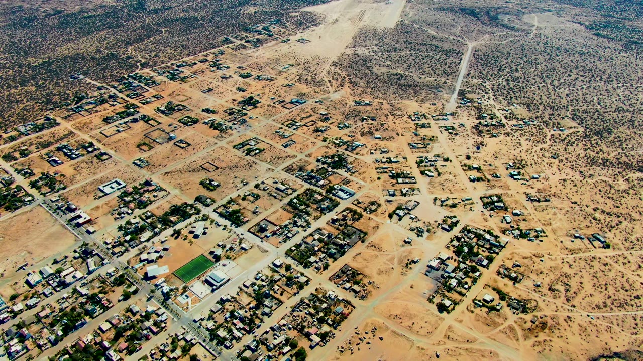 Aerial frame over San Juanico, Baja California Sur, with sandy residential blocks, scrubland, a small green field, and a pale dust plume at the top of frame, with no visible people.