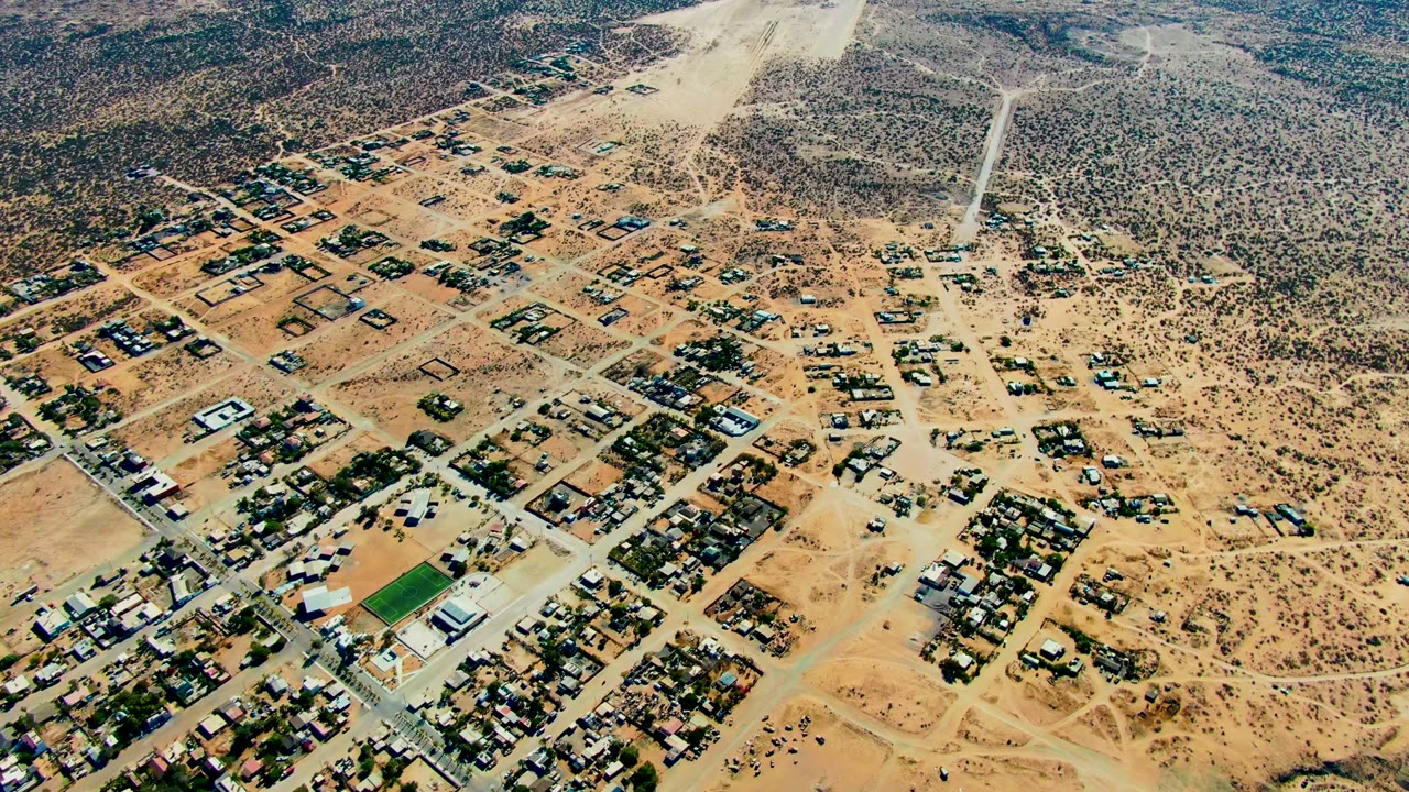 Drone view of sandy town blocks and scrub desert with dust spreading overhead.
