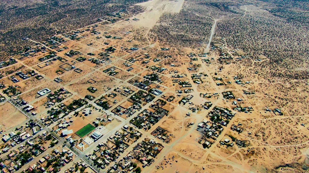 Mid-sequence aerial frame over San Juanico, Baja California Sur, with tan roads, clustered homes, open scrub, and a light dust trail crossing the upper frame, with no visible people.