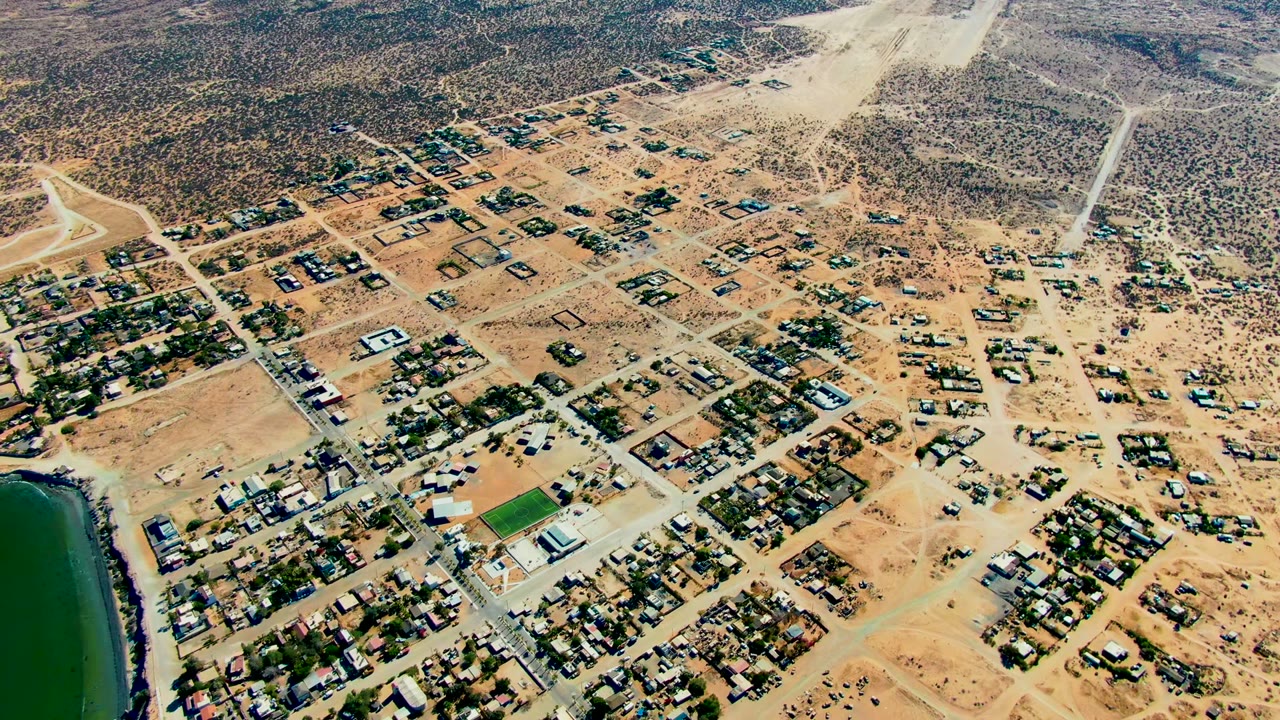 Opening aerial frame over San Juanico, Baja California Sur, showing sandy residential blocks, scattered homes, desert scrub, a green field, and a strip of water at the left edge with no visible people.