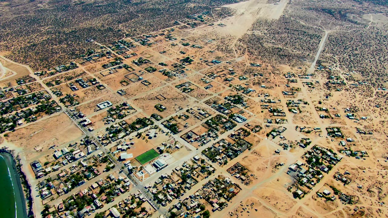 Drone frame over San Juanico, Baja California Sur, with clustered houses, tan open lots, gridded dirt streets, darker scrub in the distance, and the water edge narrowed at left, no visible people.