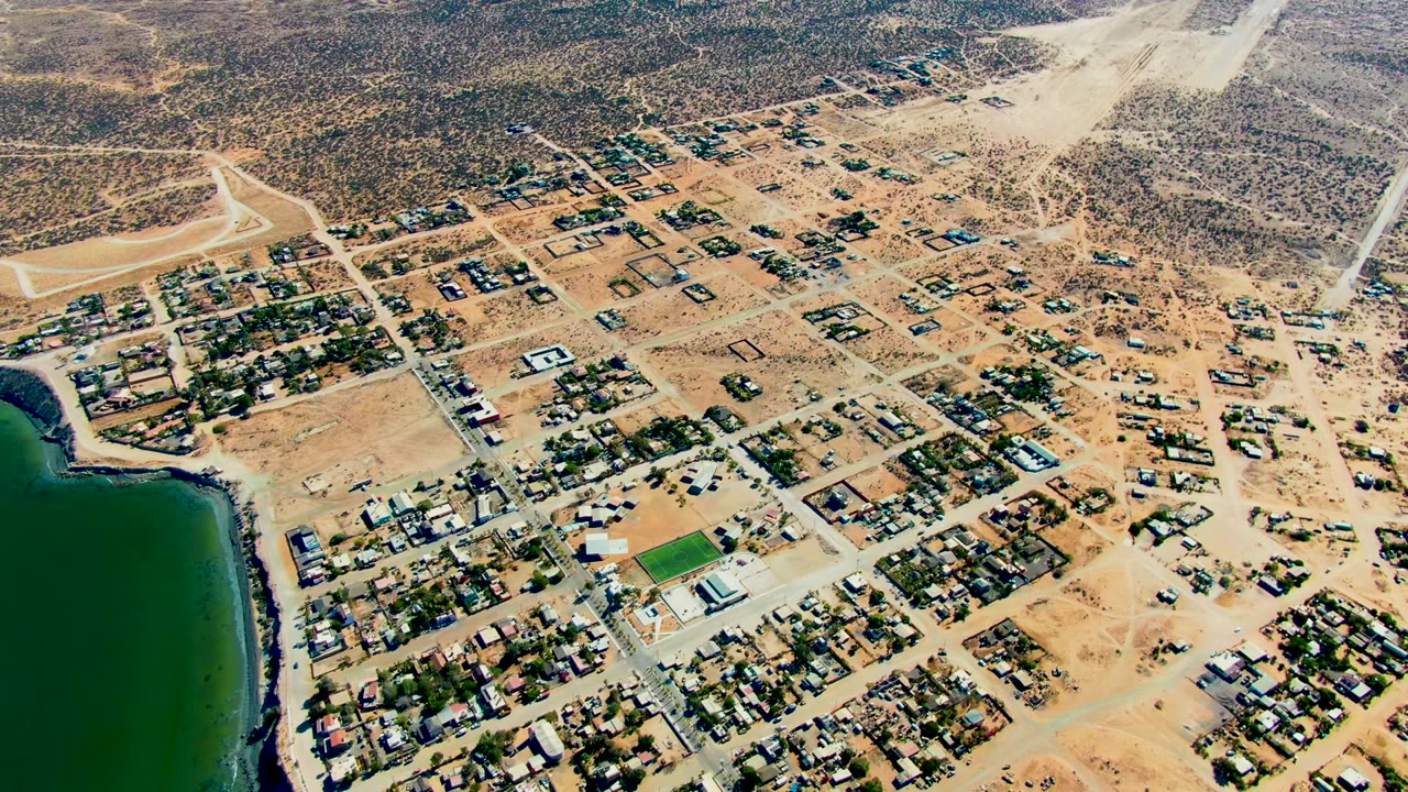 Aerial view over San Juanico, Baja California Sur, with dark green water at the left edge, dusty streets, small houses, and no visible people.
