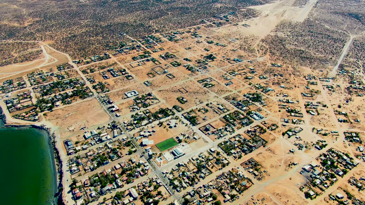 Wide aerial view over San Juanico with the shoreline water at left, small residential blocks in the center, pale dirt roads, and no visible people.