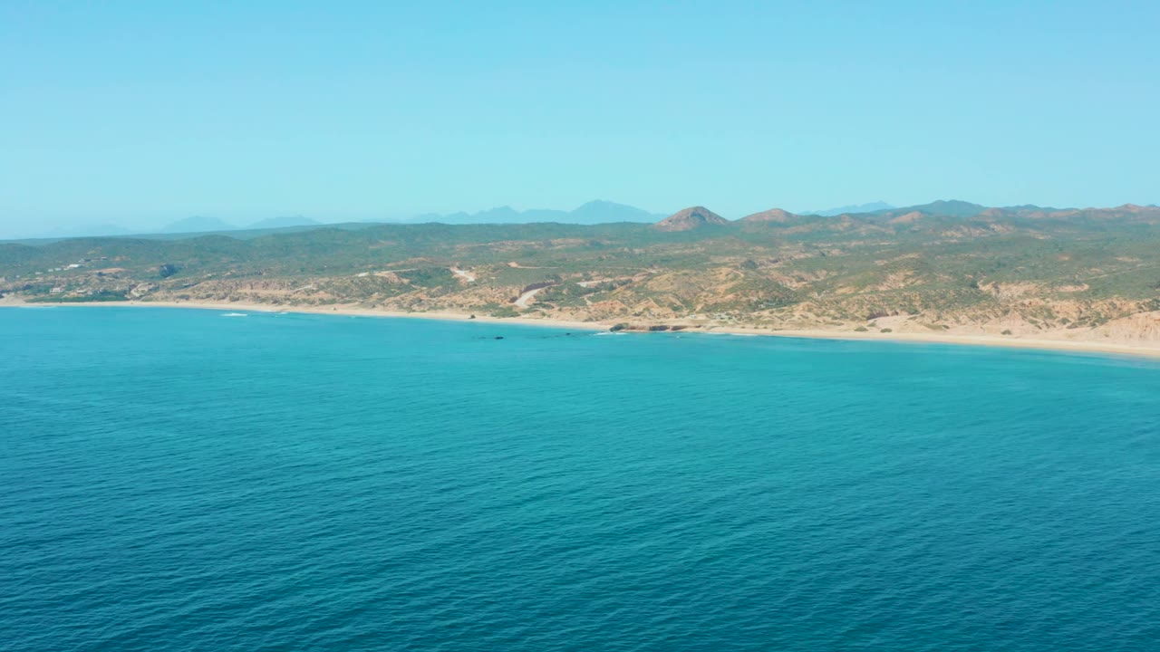 Opening aerial frame over El Tule Los Cabos with turquoise ocean in the foreground, a sandy beach across the middle, distant dry hills under clear blue sky, and no visible people.