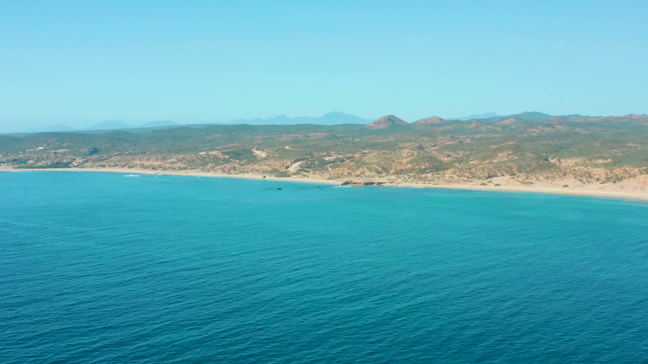 Wide coastal aerial with calm water and low hills beyond the beach