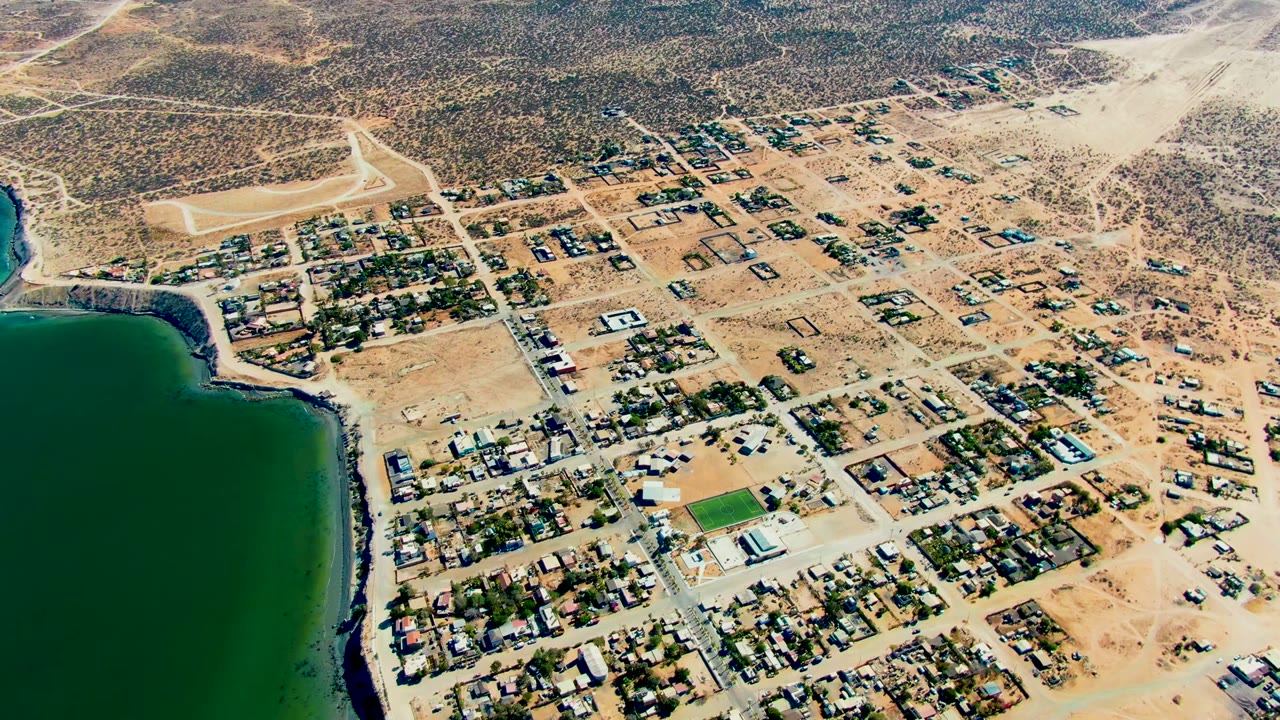 High aerial view over San Juanico, Baja California Sur, with green water along the left edge, sandy residential blocks, dirt roads, scrubland beyond, and no visible people.