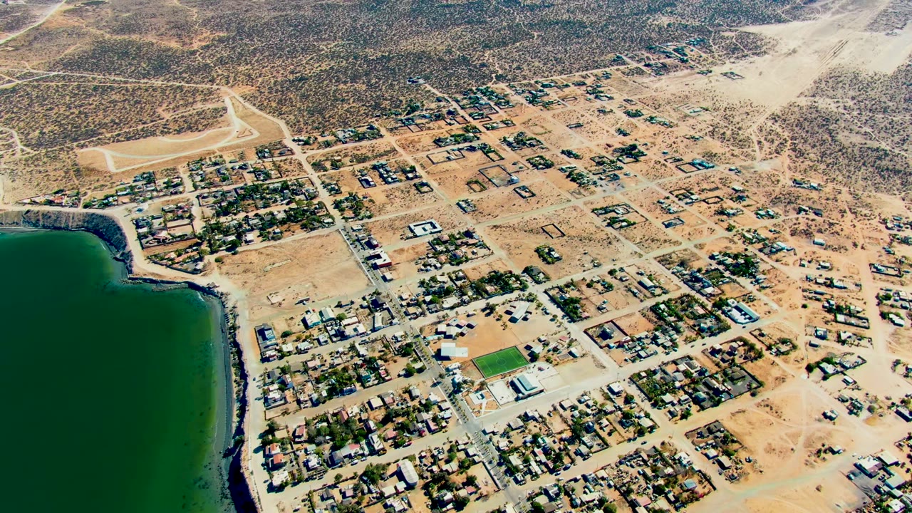High oblique aerial of San Juanico with dark green water at frame left, sandy roads and scattered homes across the center, desert tracks and scrubland in the background, and no visible people.