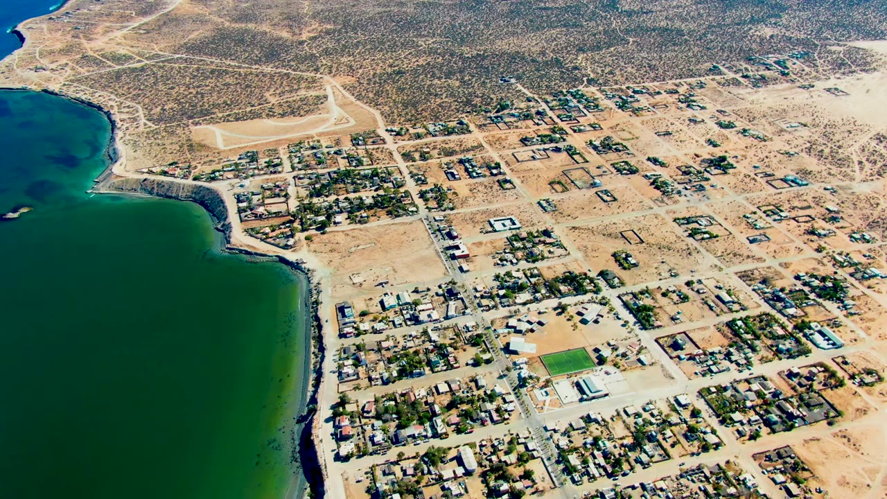 Aerial frame over San Juanico, Baja California Sur, with green-blue bay water at left, sandy streets and low homes at center, dry scrubland beyond, and no visible people.