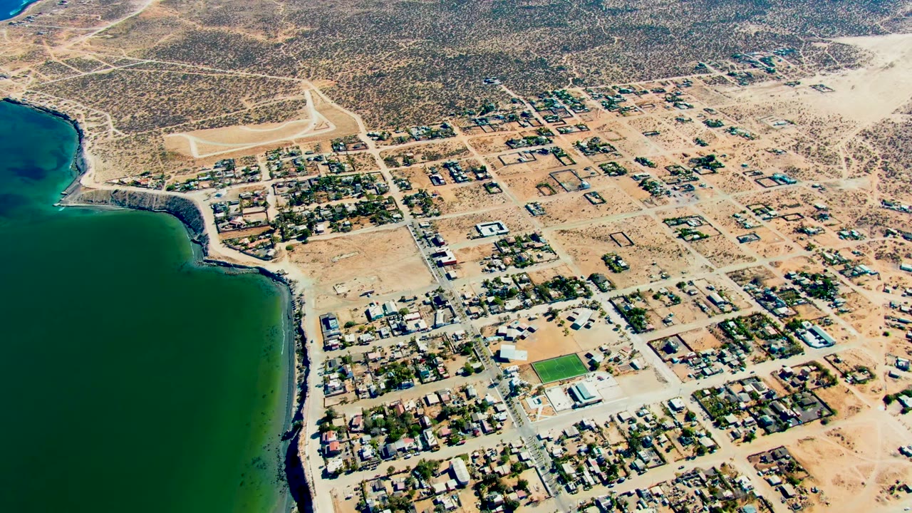 San Juanico aerial frame with dark green water along the left edge, a compact neighborhood grid below center, scattered structures to the right, dry inland brush above, and no visible people.