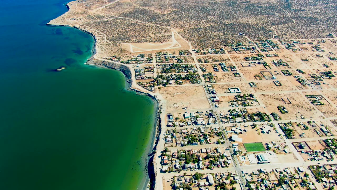 High aerial view over San Juanico, Baja California Sur, with green-blue water on the left, sandy residential blocks on the right, desert scrub beyond, and no visible people.
