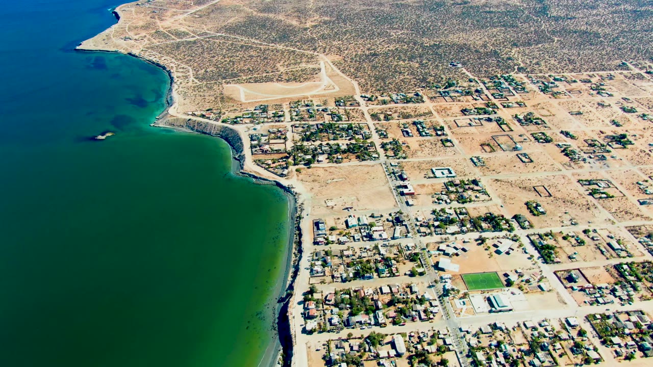Drone view of a coastal settlement beside green water and brown desert land.