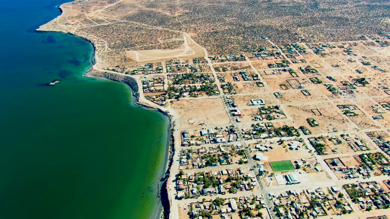 San Juanico appears from above with ocean filling the left side, a narrow rocky coast, gridded sandy streets, low buildings, a small green field, and no visible people.