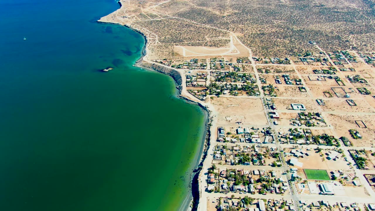 High aerial view over San Juanico, Baja California Sur, showing green-blue ocean, a sandy shoreline, residential blocks, a bright field, desert scrub, and no visible people at the start of the clip.